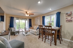 A spacious living room with a dining area, a ceiling fan, and large windows with blue curtains.
