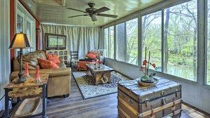 Una habitación acogedora con techo de madera, un ventanal con vistas a árboles, un sofá de cuero, un cofre de madera y una mesa con lámpara.