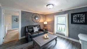 A living room with a dark leather sofa, a wooden coffee table, a floor lamp, and a large window with a view of trees. - 2nd Floor The Inn at Highland Lake (Winsted)