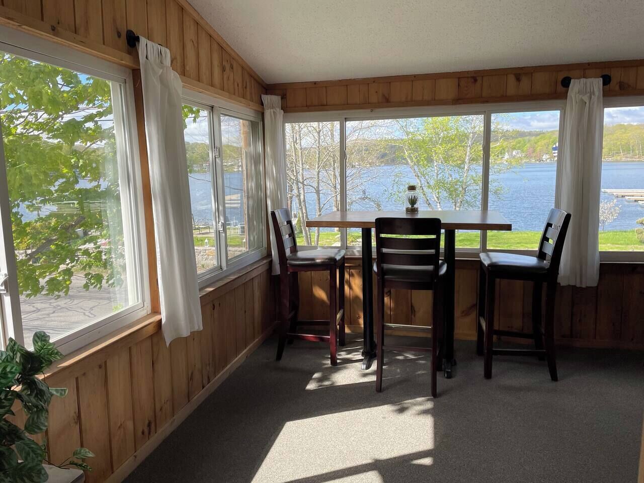 Une pièce avec un mur lambrissé en bois, une table entourée de quatre chaises et de grandes fenêtres donnant sur une étendue d’eau.
