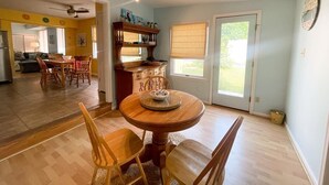 A dining area with a round wooden table and chairs, a kitchen in the background, and a large window with blinds. - Walk to Washington St. Mall (West Cape May)