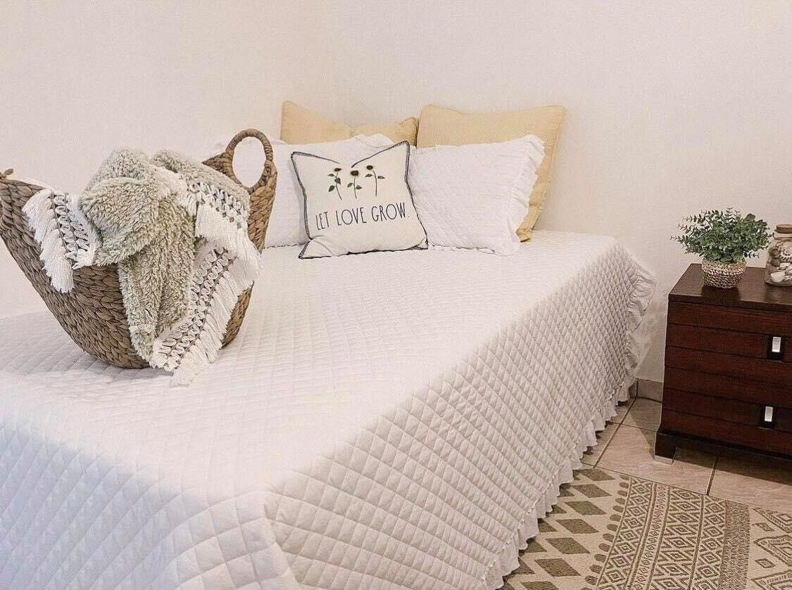 A neatly made bed with a white quilt, a woven basket with a blanket, and a pillow with the text "LET LOVE GROW."