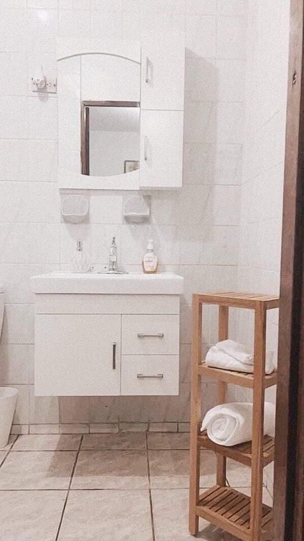 A bathroom with a white sink, mirror, and cabinet, a wooden shelf with towels, and tiled flooring.