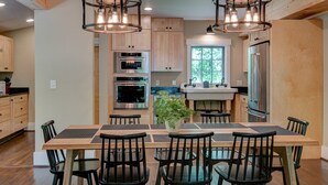 A modern kitchen with wooden cabinets, stainless steel appliances, a central island, and black chairs. - Orchard House - creekfront with outdoor fireplace, hot tub, hiking on site (Banner Elk)
