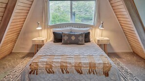 Een slaapkamer met een groot bed, twee bedlampen, een raam met uitzicht op groen, en een schuine plafond.