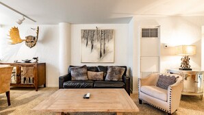 Ein Wohnzimmer mit Couchtisch aus Holz, einer schwarzen Ledersofa mit Zierkissen, einem beigen Sessel, einem Holznachttisch und einem an der Wand befestigten Damhirschschädel.