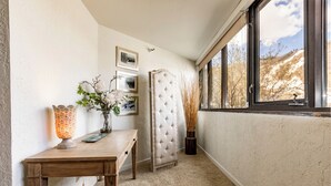 Ein Eckzimmer mit Holztisch, weißem Sitzbänkchen, Blumen in einer Vase und einem Fenster mit Blick auf Bäume und Hügel.