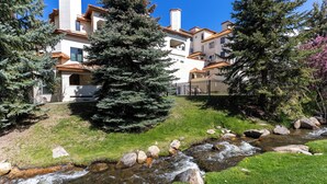 Un edificio de varios pisos con fachada de piedra, un área verde con un arroyo y un gran árbol de hoja perenne.