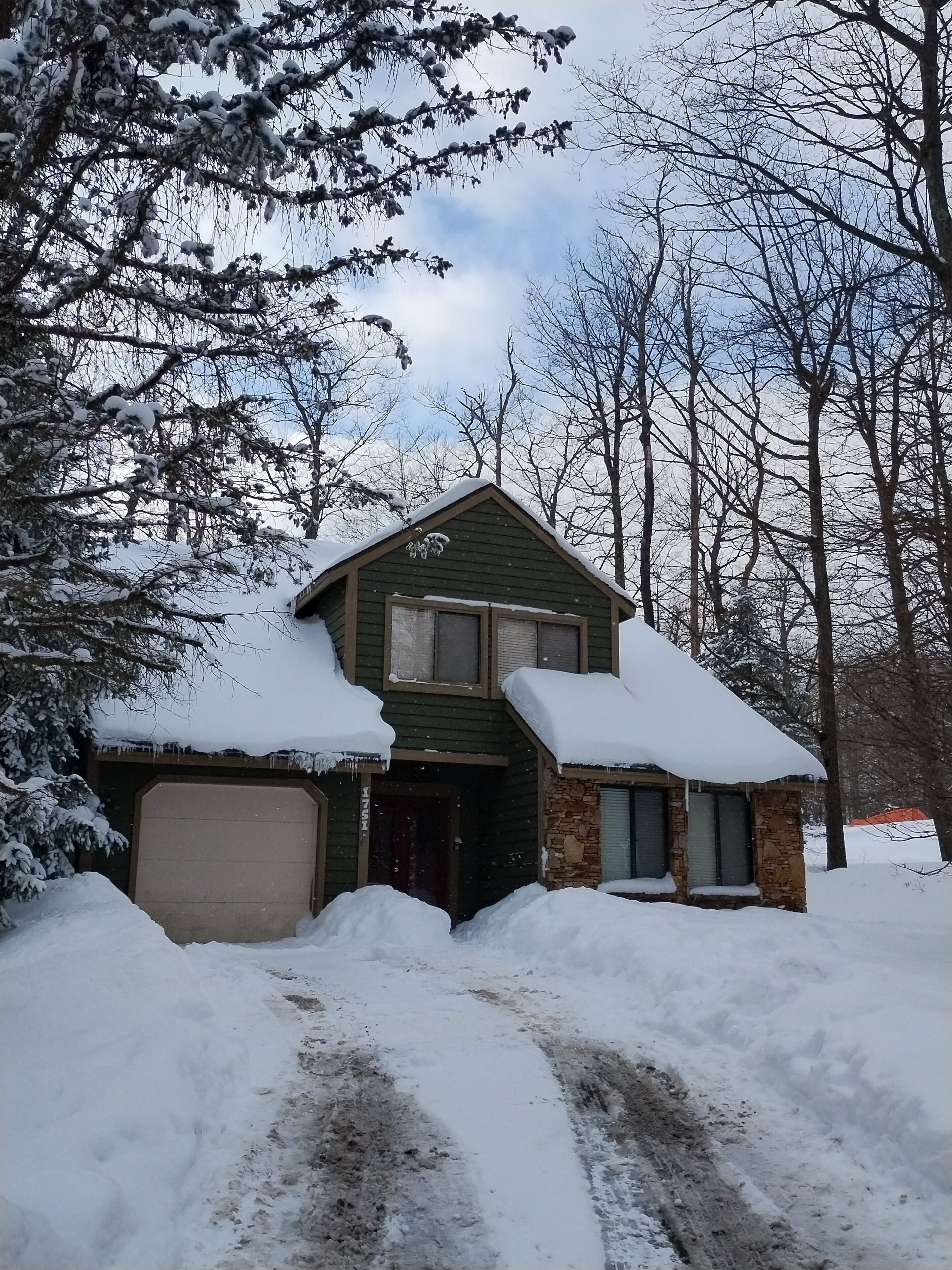 A snow-covered house with a garage, a stone foundation, and a driveway.