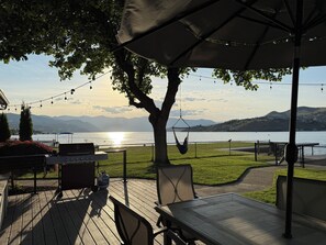 A patio with a table and chairs, a hanging chair, a grill, and a view of a lake with mountains in the background.
