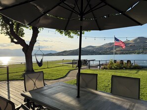 An outdoor patio with a table and chairs, a hanging chair, a fence, and a view of a lake and mountains.
