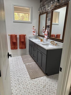A bathroom with a white tiled floor, a window, two sinks, a mirror, and two towels hanging on a towel rack.