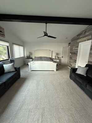 A bedroom with a bed, two nightstands, a ceiling fan, a window with blinds, a dark-colored sofa, and a wall with a wooden panel.
