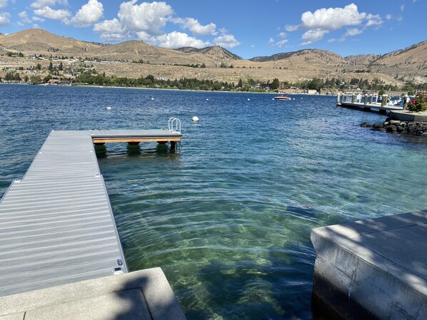 Un lago cristalino con un muelle que se adentra en el agua, rodeado de montañas y un cielo despejado.