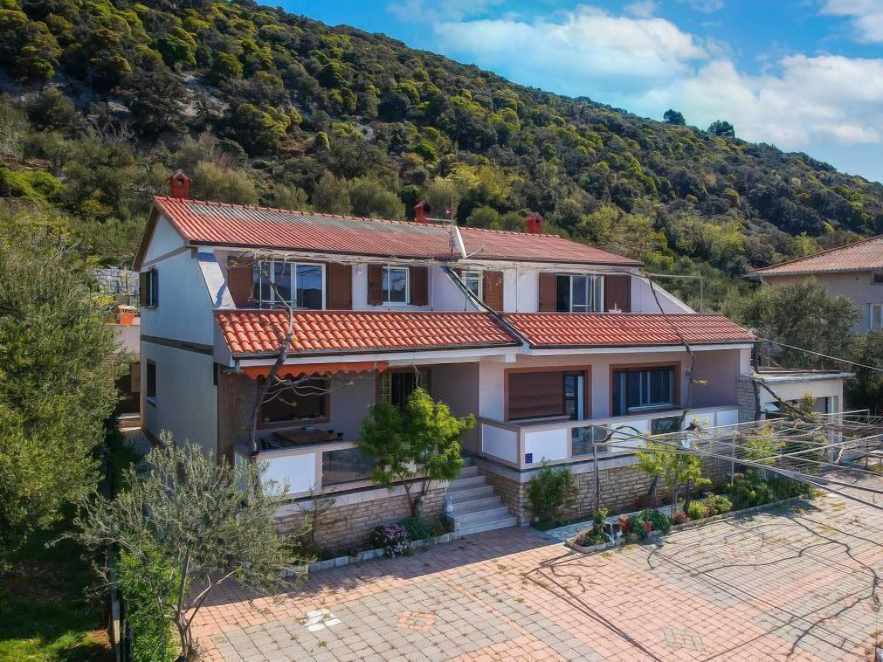 A two-story house with a red-tiled roof, surrounded by greenery and hills.