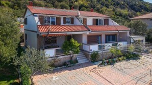 Uma casa de dois andares com telhado de telhas vermelhas, rodeada por vegetação e colinas.