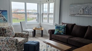 Un salon avec un canapé marron, un fauteuil à motifs et un pouf, une table basse et un tableau accroché au mur.
