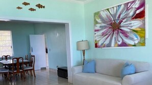 A living room with a sofa, coffee table, and a painting. There is a dining area with chairs and a table in the background. - Situated on the Ocean Jacaranda Bay Villa  (Nassau)