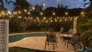 A poolside area with string lights, chairs, and a table. - Situated on the Ocean Jacaranda Bay Villa  (Nassau)