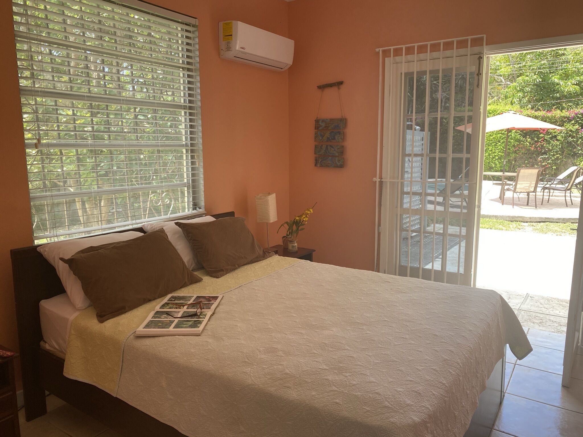 Une chambre avec un lit, une fenêtre avec des stores, un climatiseur, une table de chevet avec une lampe et une vue sur une terrasse extérieure.