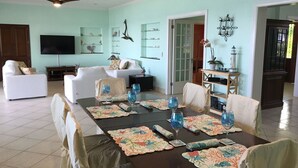A dining area with a table set for six, surrounded by chairs. There is a television mounted on the wall, a sofa, and a cabinet with decorative items. - Situated on the Ocean Jacaranda Bay Villa  (Nassau)