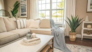 Ett vardagsrum med en beige soffa, ett soffbord med böcker och en tekanna, en stor växt och en mönstrad matta.