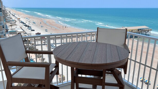A balcony with a wooden table and chairs overlooking a beach with a pier. - Luxury Oceanfront Condo in Daytona Beach Shores with Gorgeous Views! 4BR, 3BA (Daytona Beach Shores)