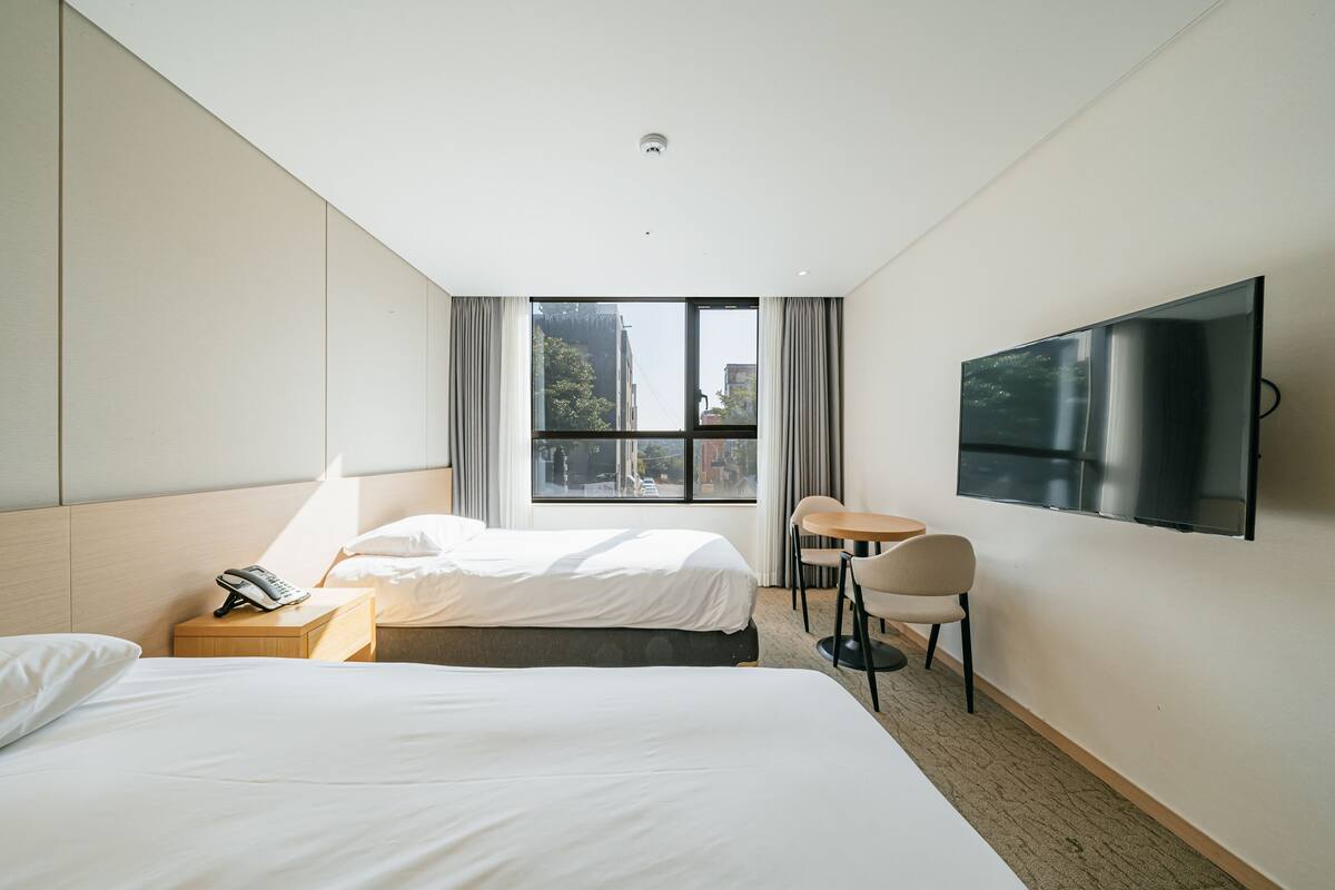 A hotel room with two beds, a desk, a chair, a TV, and a window with a view of buildings and trees.