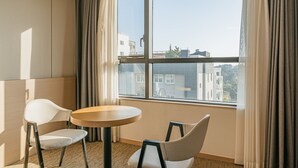 A hotel room with a large window, two chairs, a round table, and a view of buildings outside. - Hotel ZINC (Seogwipo)