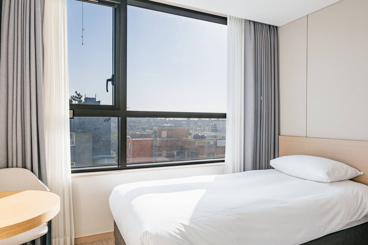 A hotel room with a large window, a bed, a bedside table, and a view of the cityscape.