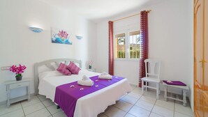 A bedroom with a bed, a chair, a nightstand, a window with curtains, and a wall with a painting. - 0306 Villa Monte Verde Granada (Javea / Xàbia)