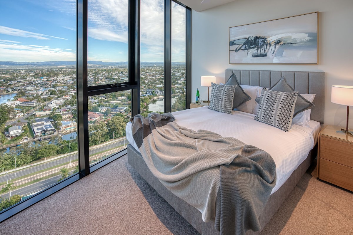 A modern bedroom with a large bed, a view of a cityscape, and a painting on the wall.