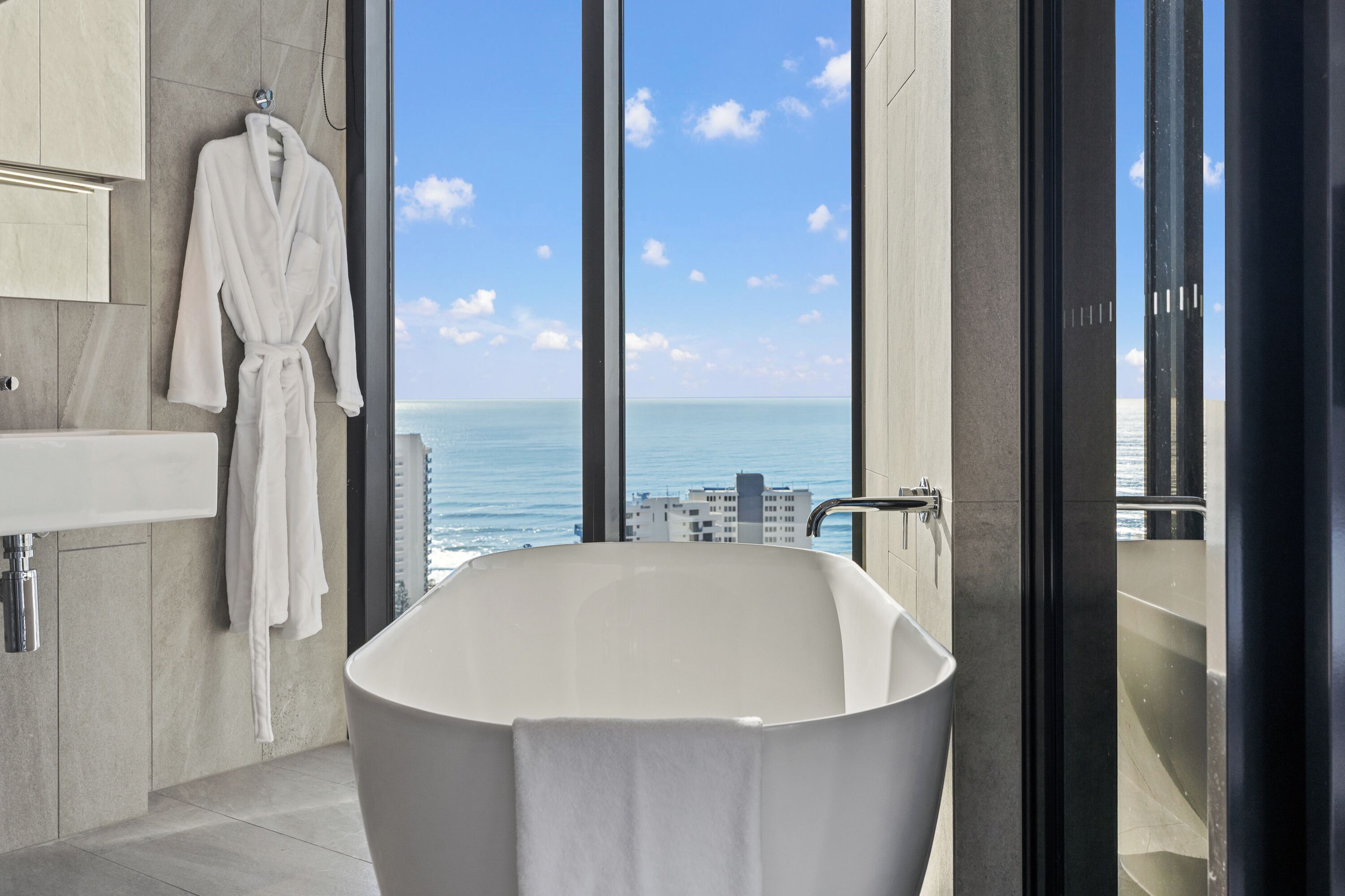 A modern bathroom with a freestanding bathtub, a large window with a view of the ocean, and a robe hanging on the wall.