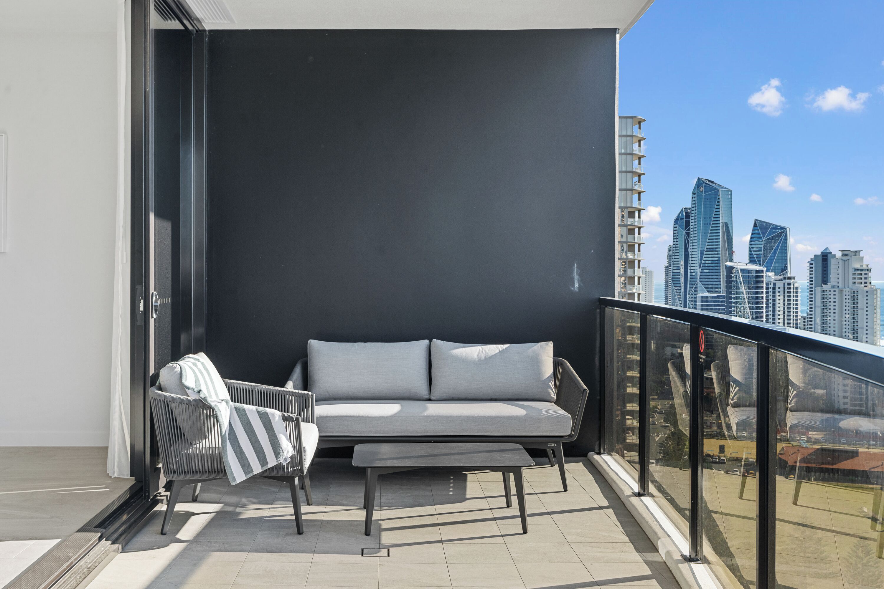 Ein Balkon mit einer grauen Couch und einem gestreiften Sessel, einer Glastür und Blick auf eine Stadtlandschaft.
