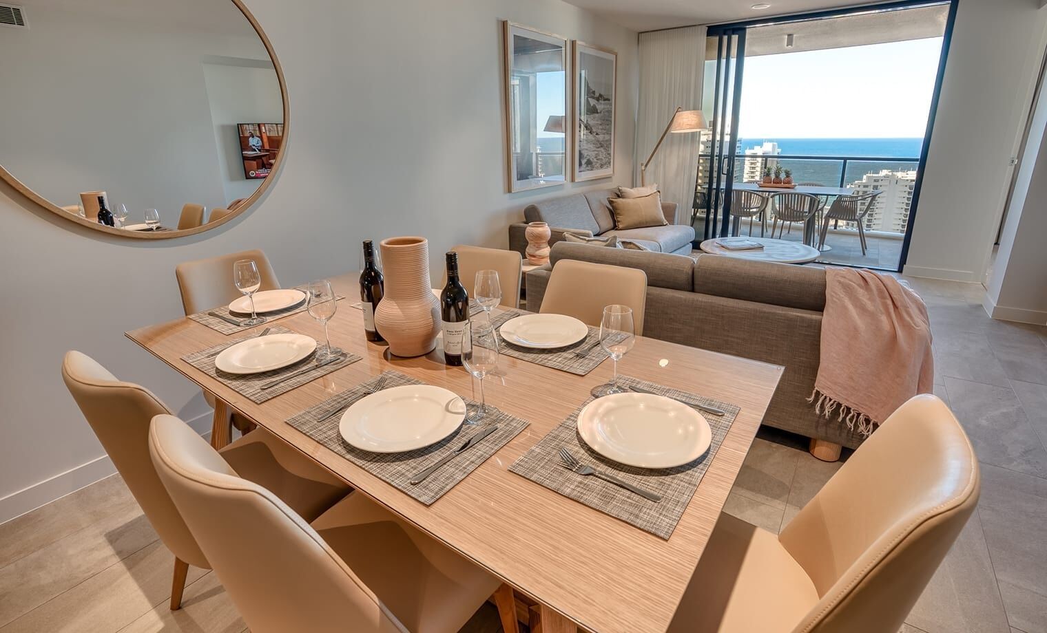 A dining area with a wooden table set for six, surrounded by beige chairs. A large mirror and framed artwork are on the wall.
