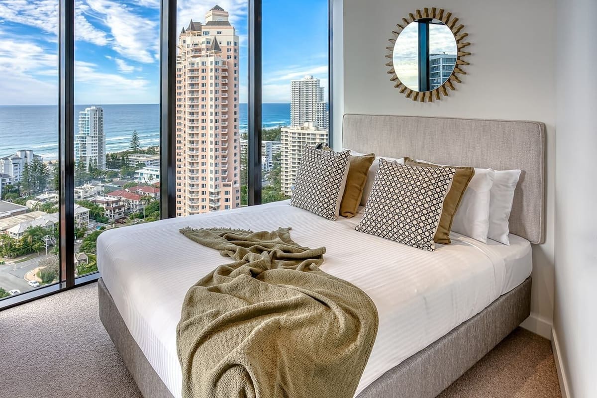 A hotel room with a large bed, a view of the cityscape, and a sunburst mirror on the wall.