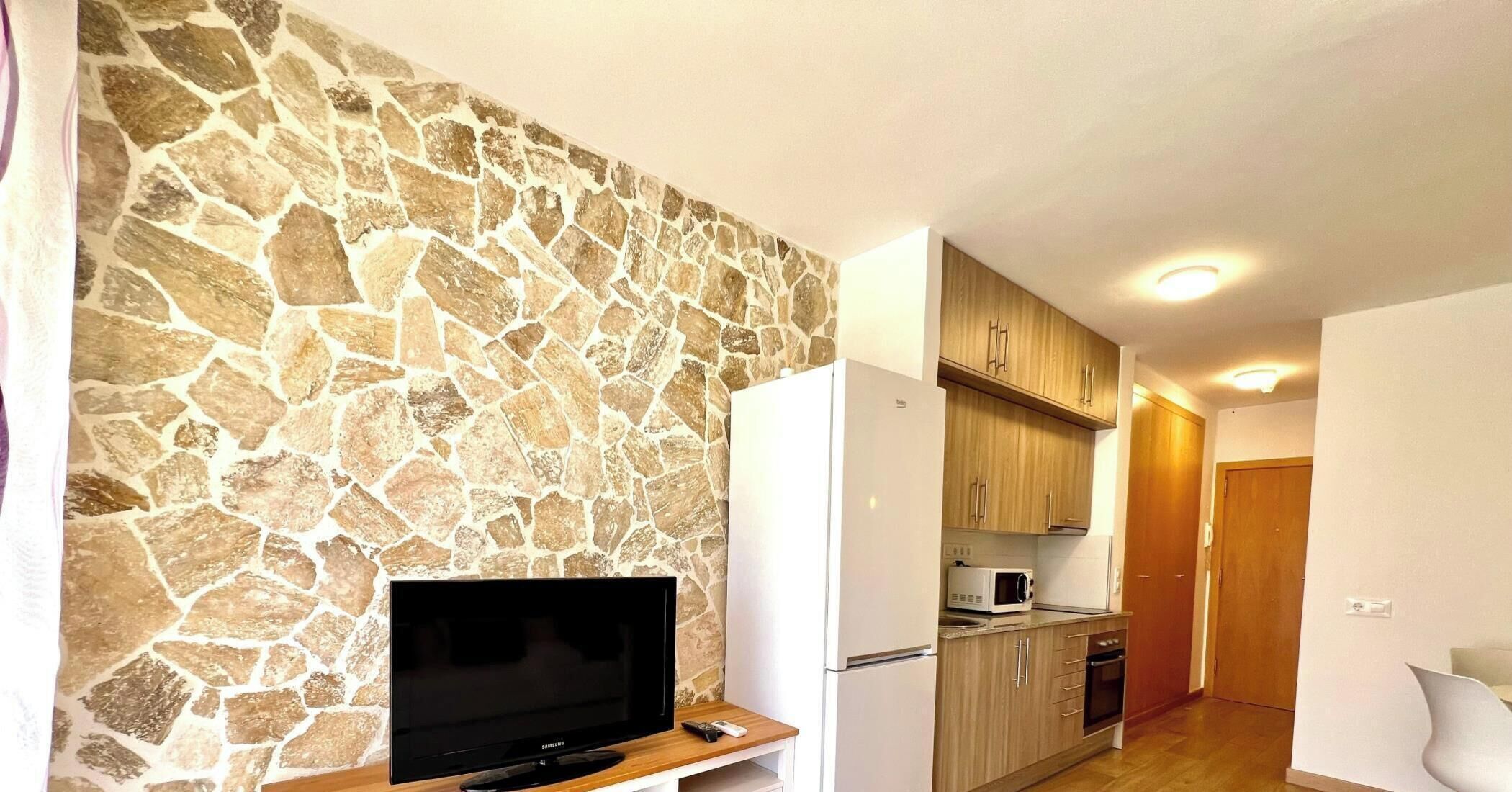 Een moderne keuken met een stenen muur, houten kasten, een inbouwmagnetron en een tv op een houten standaard.