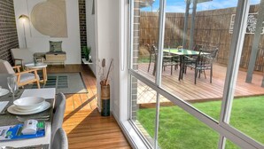 A modern interior with a dining area, a living room, and a view of a wooden deck and garden. - Sleek Stylish Modern Close to Airport (Fraser Rise)
