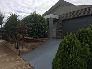 A modern building with a garage door, a concrete driveway, and landscaped areas with various plants. - Sleek Stylish Modern Close to Airport (Fraser Rise)