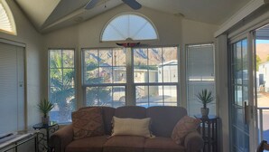 Ein Wintergarten mit Deckenventilator, einem großen Fenster mit Jalousien, einem braunen Sofa mit gemusterten Kissen und einem kleinen Tisch mit einem Blumentopf.