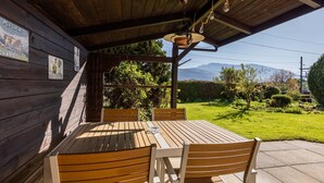 Um alpendre coberto com mesa e cadeiras de madeira, um lustre pendurado e vista para uma paisagem verde com montanhas ao longe.