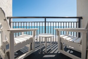 Een balkon met twee witte Adirondack-stoelen en een tafeltje, met uitzicht op een strand met helderblauw water.