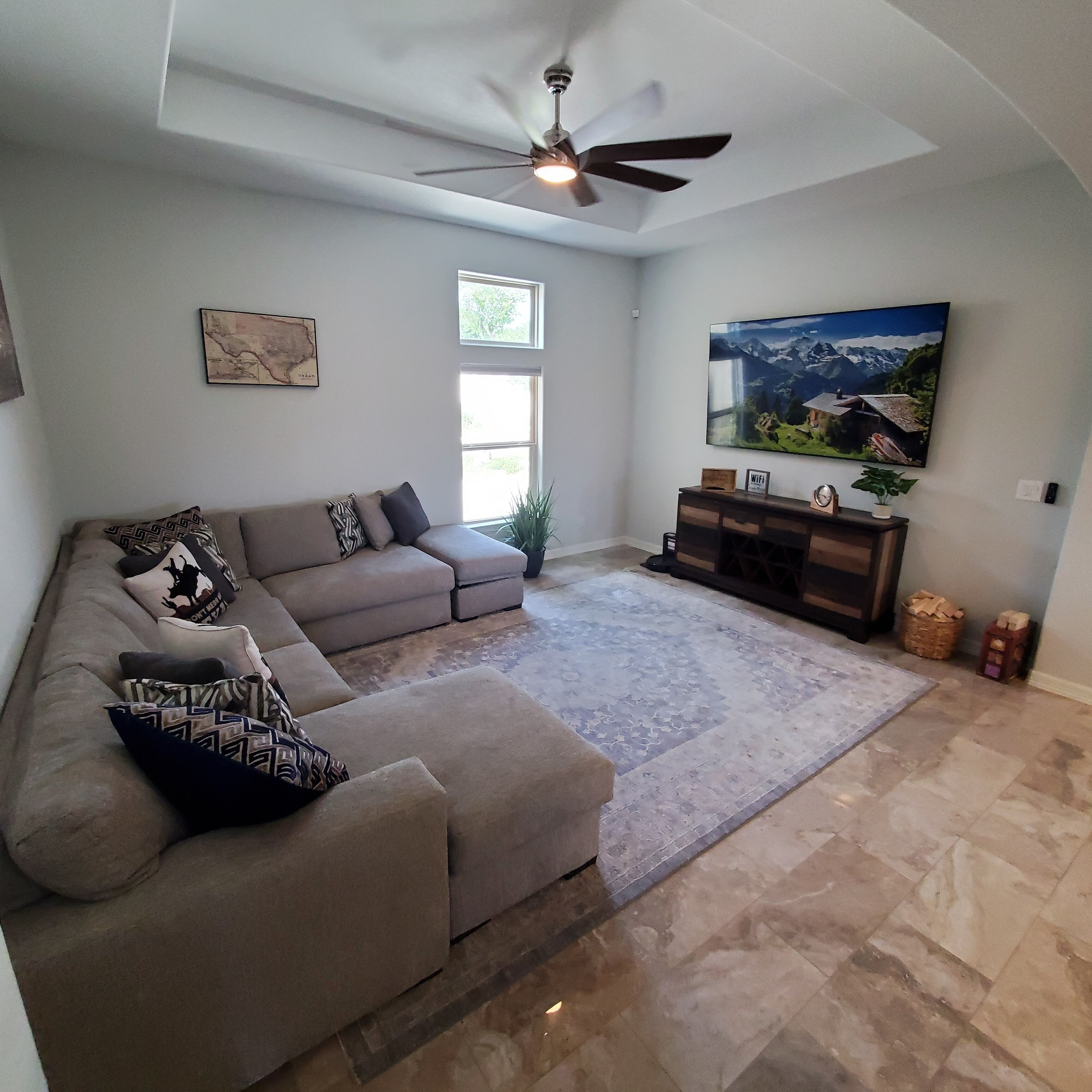 A living room with a sectional sofa, a ceiling fan, a large window, a painting, and a small wooden cabinet.