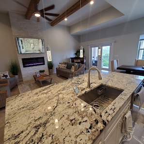 A spacious kitchen with a large island, a built-in oven, and a sink. There is a dining area with a table and chairs, and a living room with a sofa and a fireplace.