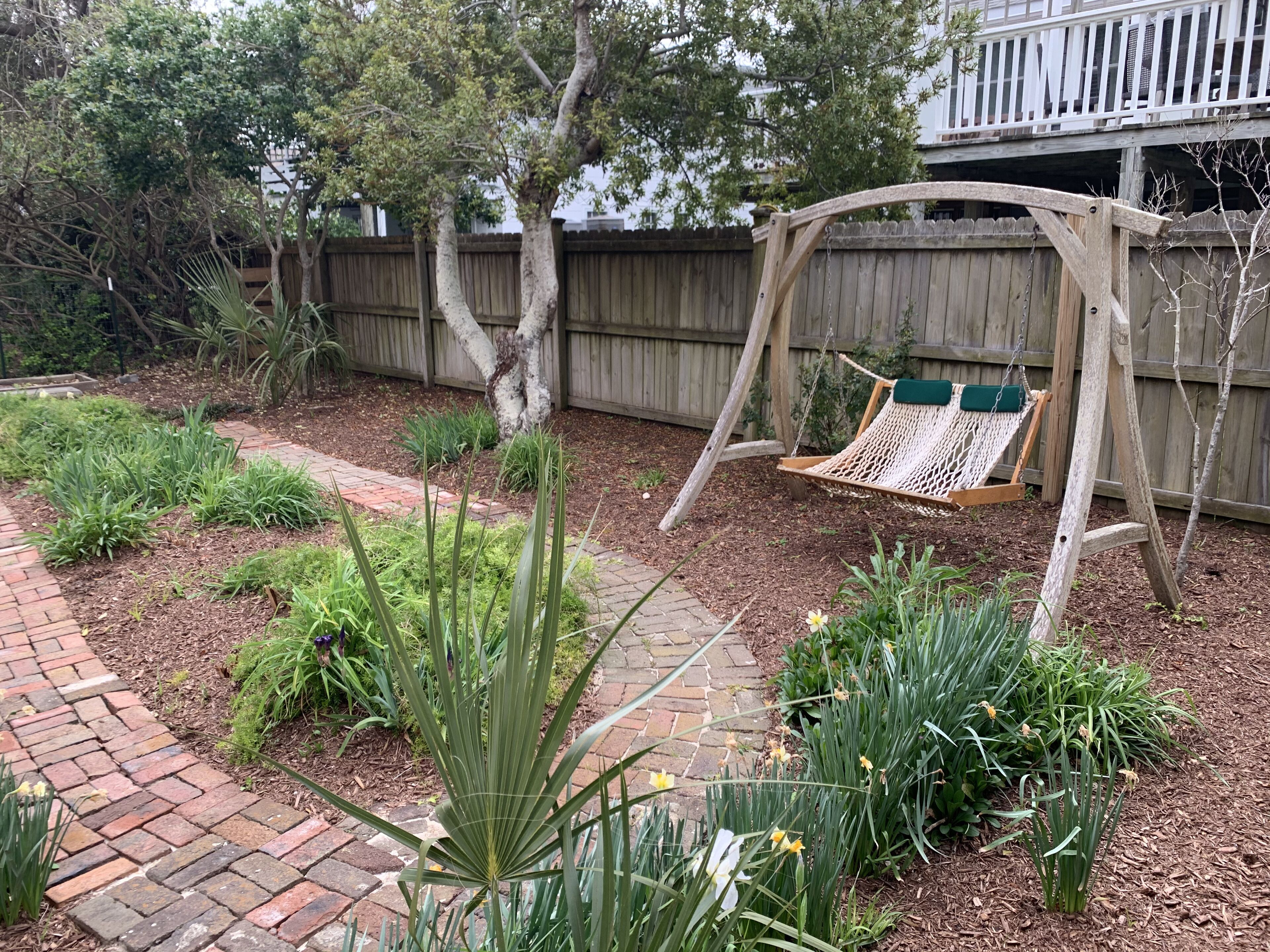 Un jardín trasero con un conjunto de columpios de madera, un sendero de ladrillo y diversas plantas, incluyendo narcisos.