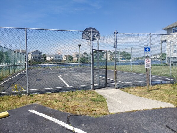 An outdoor basketball court enclosed by a chain-link fence with a basketball hoop, a no dogs allowed sign, and a parking area with a yellow bollard. - Private Home (Kill Devil Hills)