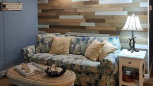 Ett vardagsrum med en mönstrad soffa, en ottoman i rotting, ett sidobord med en lampa och en vägg med en träpaneldesign.