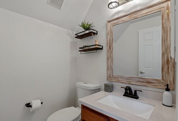 A bathroom with a toilet, sink, mirror, and a small shelf with a plant.