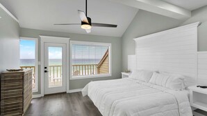 Un dormitorio con cama, ventilador de techo, mesita de noche, ventana con persianas y vista al océano.