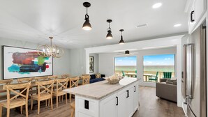 Una cocina moderna con una isla grande, sillas de madera, iluminación colgante y vista al océano.